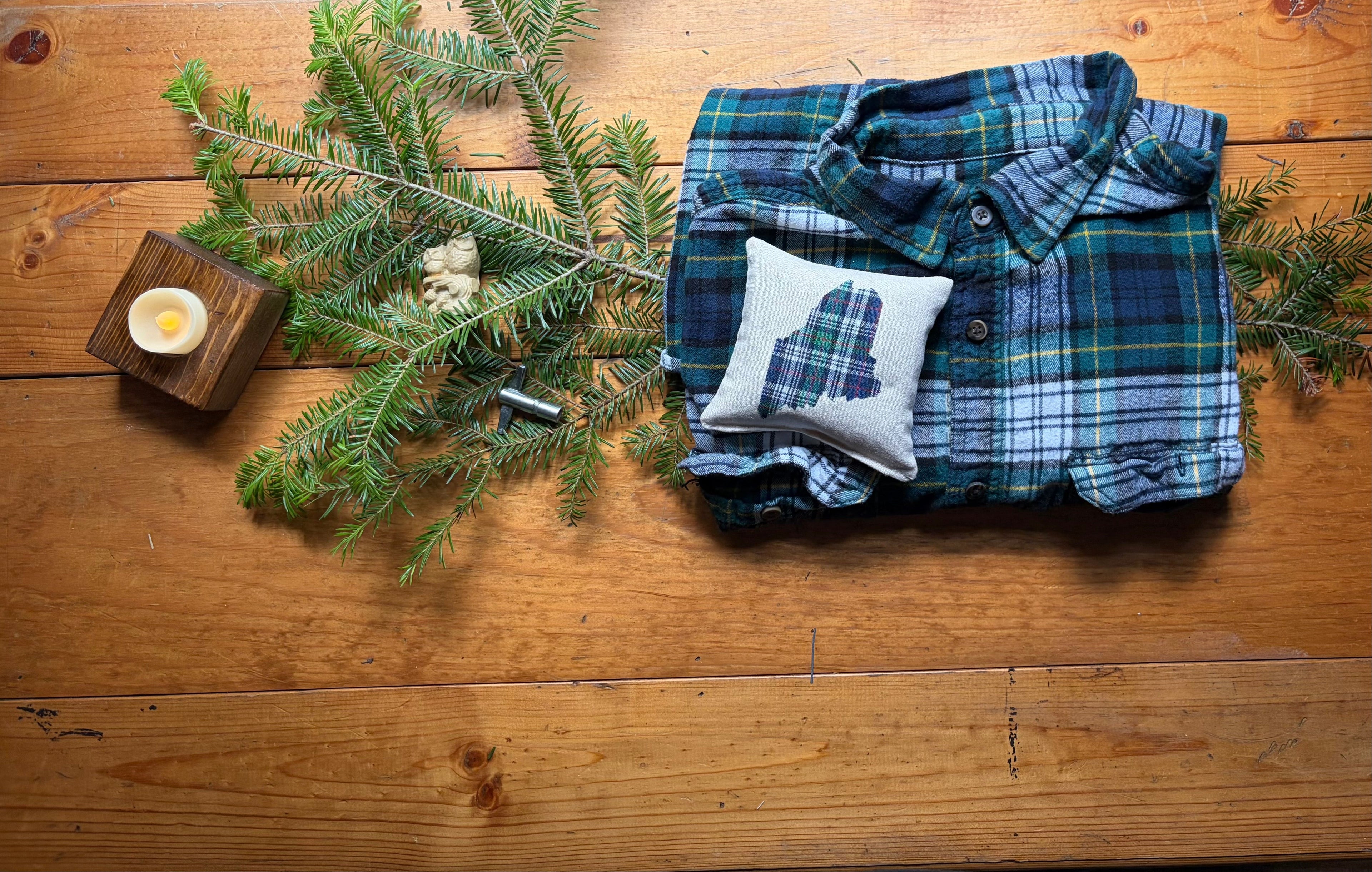 Flannel, Fir & Firelight collection featuring a folded flannel shirt, handmade Dad's Plaids Maine Balsam Fir sachet by Perry Home Naturals, fresh balsam fir branches, wooden tea light holder, and family keepsakes on a rustic wooden table.
